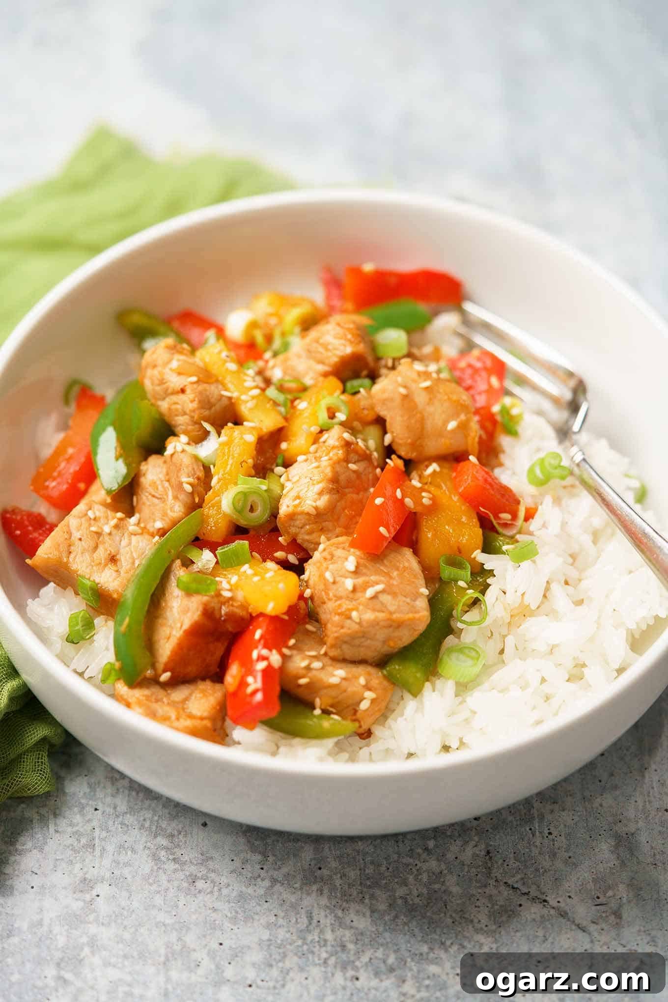 A single serving bowl of homemade sweet and sour pork, featuring tender pork pieces, vibrant red and green bell peppers, and juicy pineapple, served over a bed of rice and garnished with fresh scallions.