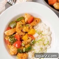A bowl of sweet and sour chicken with pineapple and red, green, and yellow bell peppers next to a serving of rice and garnished with sesame seeds and scallions.