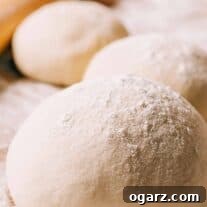 Round balls of pizza dough on a cutting board, dusted with flour, a rolling pin in the background.