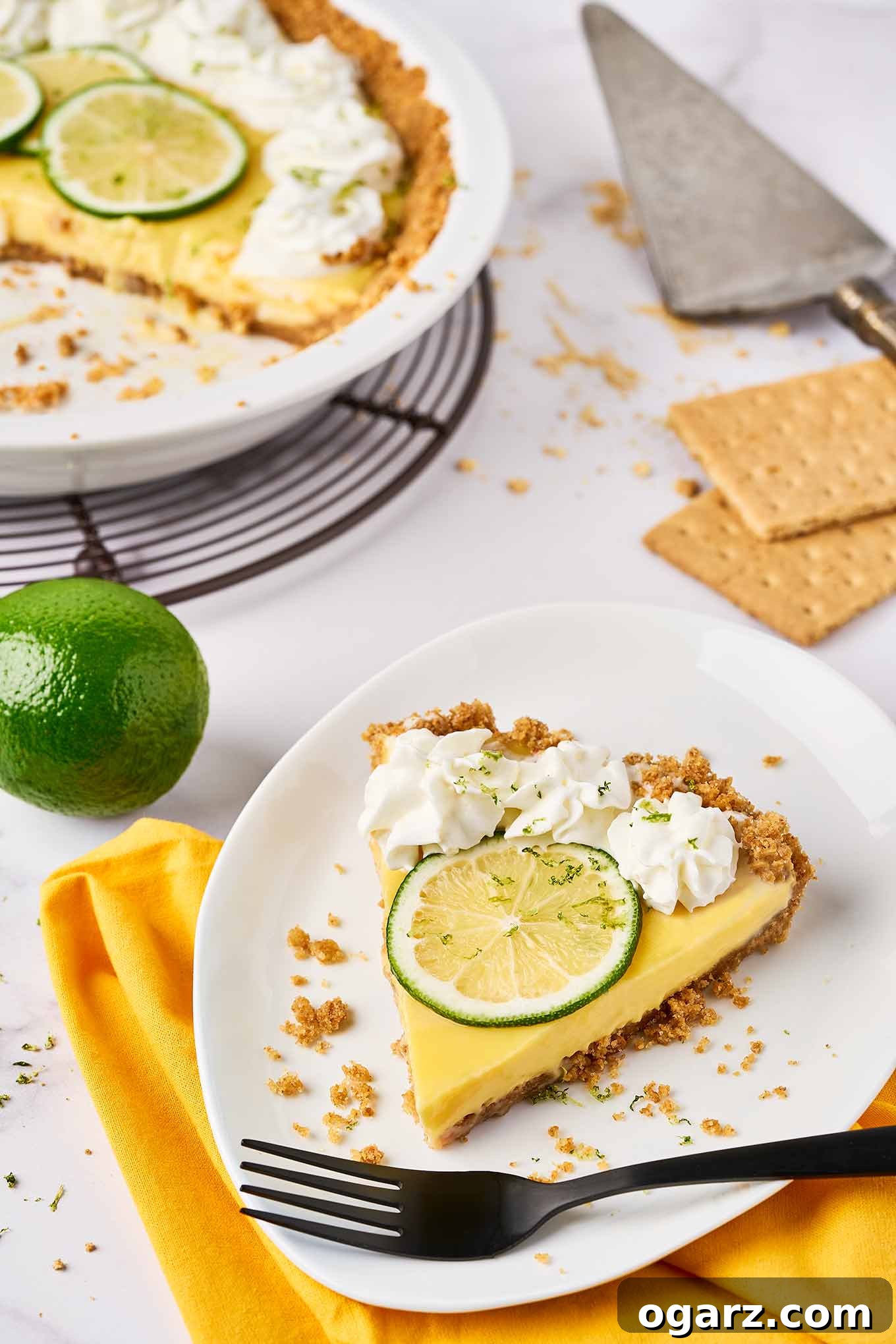 Sunshine State Key Lime Pie 6 A white dessert plate with a single slice of key lime pie and a black fork with the remaining pie and a pie server in the background.