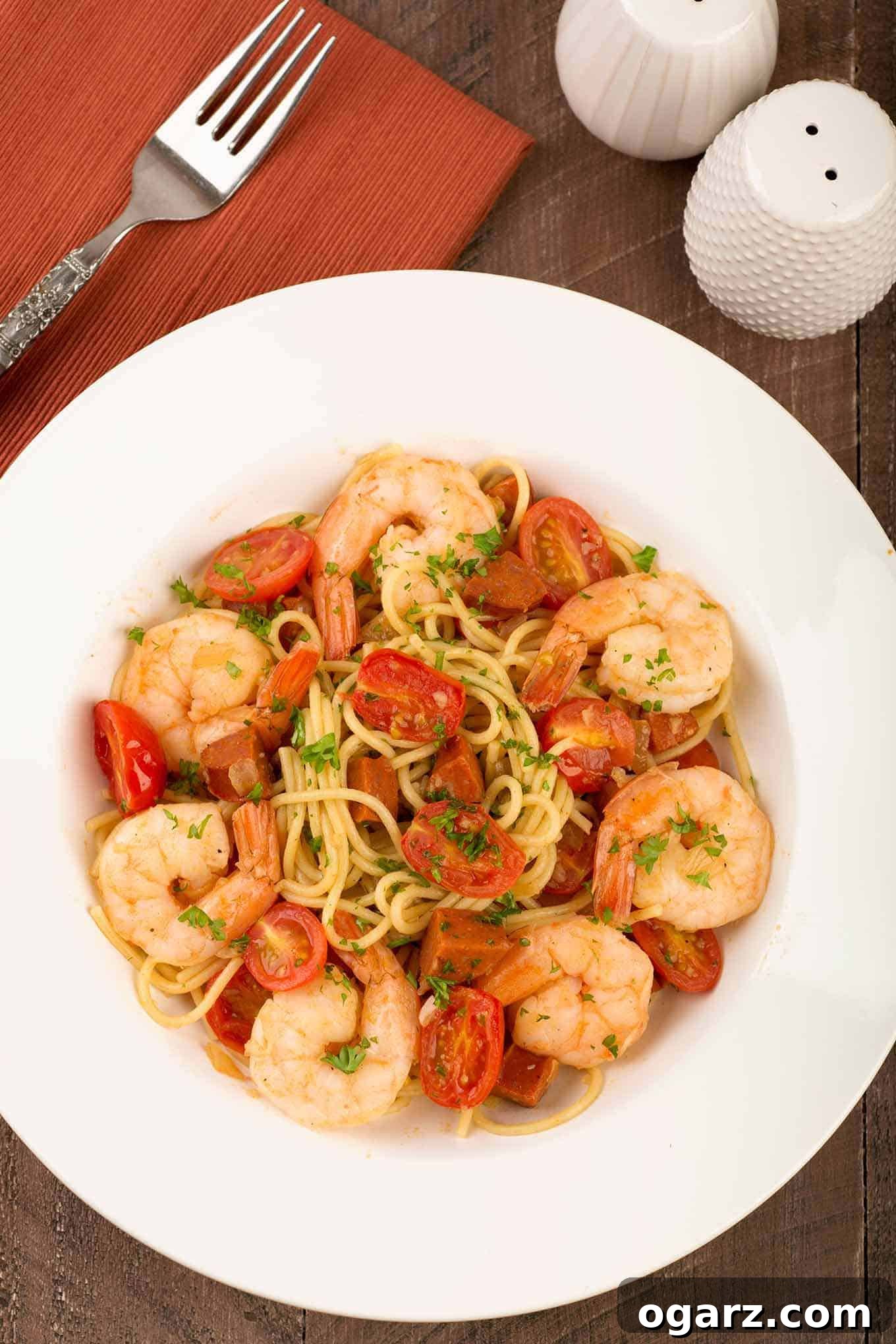 An overhead view of a serving of thin spaghetti tossed with chunks of chorizo, sautéed garlic and onion, jumbo shrimp simmered in sherry, and grape tomatoes. 
