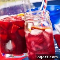 A mason jar filled with summer berry sangria with a red and white striped straw in it, a pitcher with more sangria with fruit in it in the background.