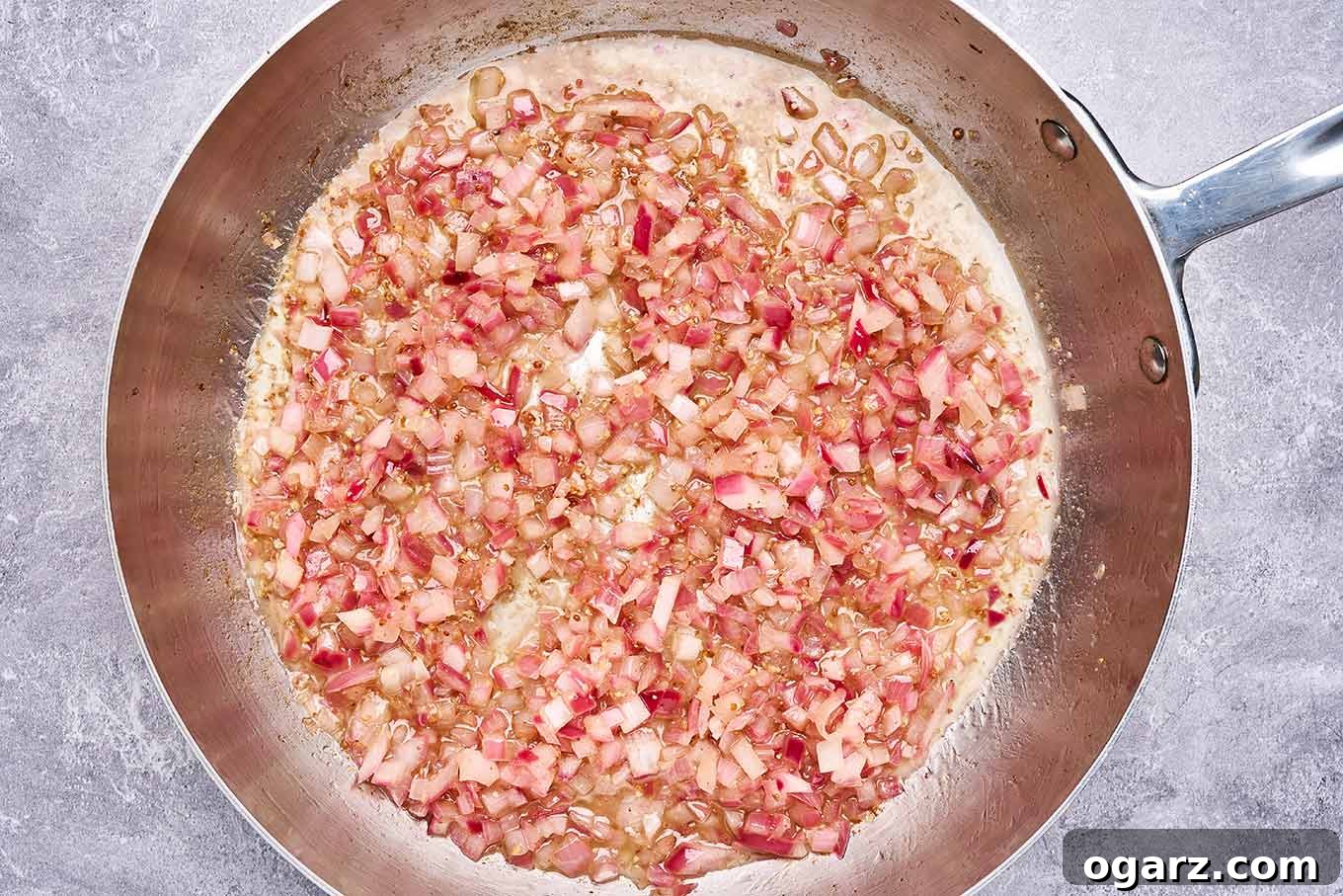 Authentic Warm German Potato Salad 4 Preparation step for warm bacon dressing featuring an overhead view of a stainless frying pan with diced red onion sautéed in oil and bacon drippings.