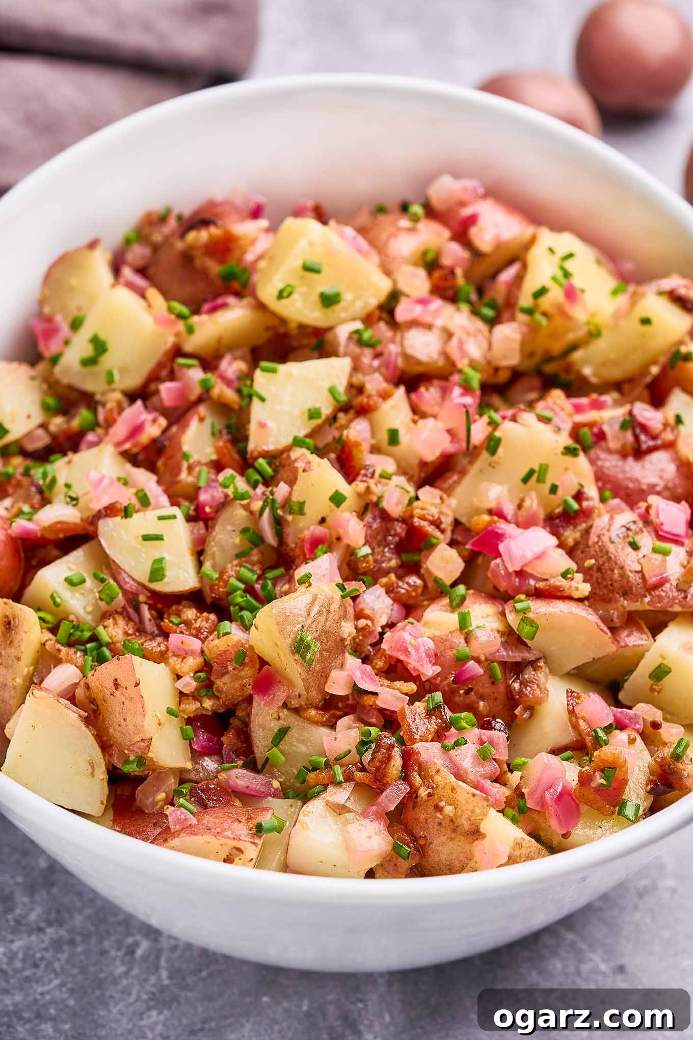 Authentic Warm German Potato Salad 5 A white oval serving bowl filled with warm German potato salad on a gray concrete surface.