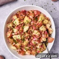A serving bowl filled with warm potato salad with bacon dressing, serving spoon to the side, surrounded by fresh ingredients and scattered chopped chives.