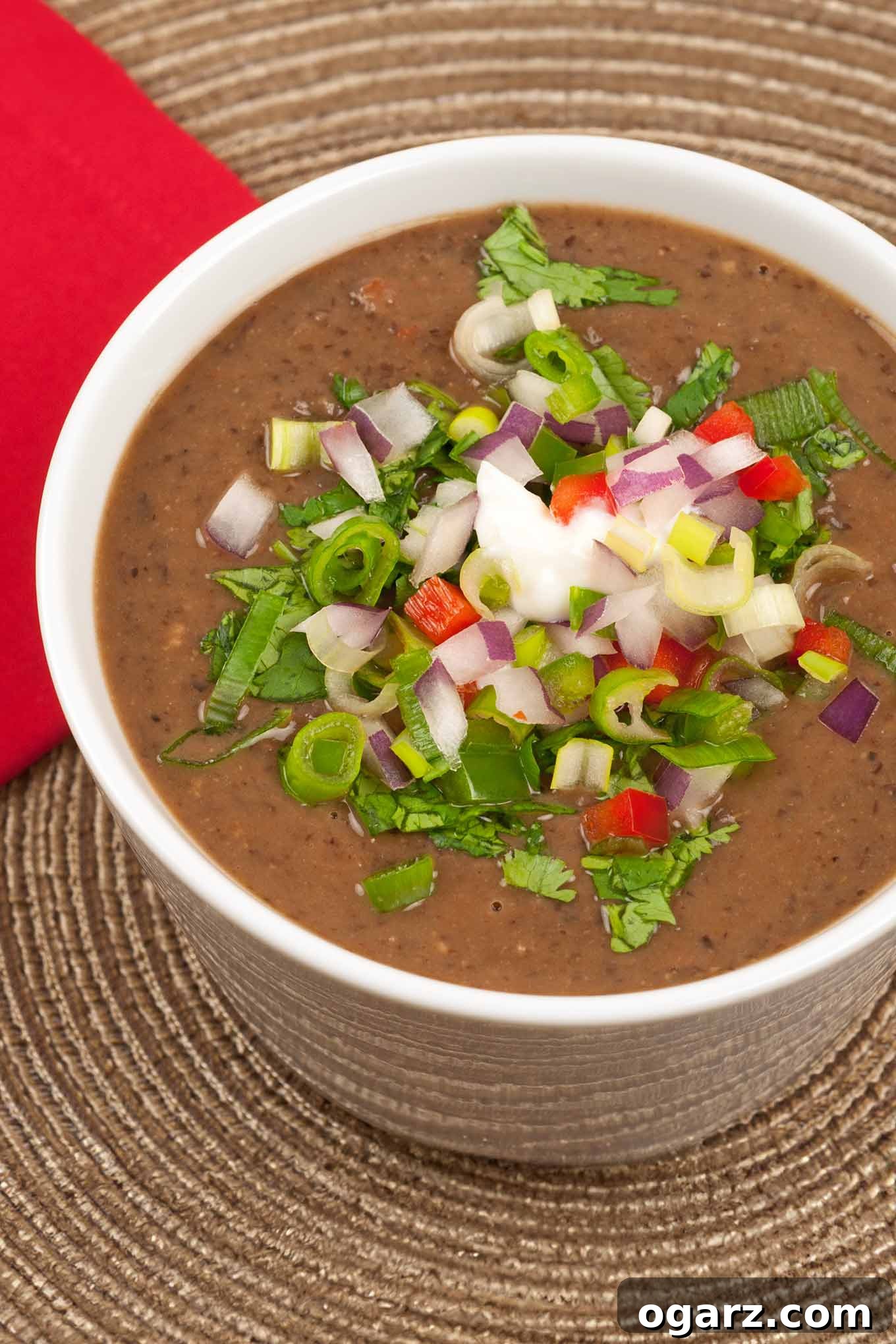 A close-up photo of a cup of black bean soup topped with diced fresh vegetables including jalapeno and red bell peppers, red onion, scallions, and cilantro on a rattan placemat. A vibrant, fresh garnish adds visual appeal and a burst of flavor to this hearty and healthy soup.