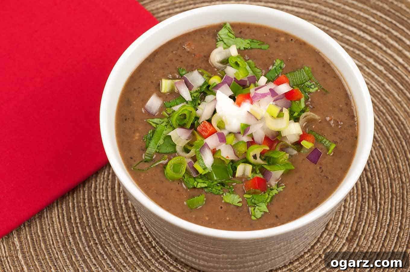 A close-up photo of a cup of black bean soup topped with diced fresh vegetables including jalapeno and red bell peppers, red onion, scallions, and cilantro on a rattan placemat. A perfect harmony of color, texture, and taste in every spoonful.