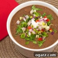 A close-up photo of a cup of black bean soup topped with diced fresh vegetables including jalapeno and red bell peppers, red onion, scallions, and cilantro on a rattan placemat.
