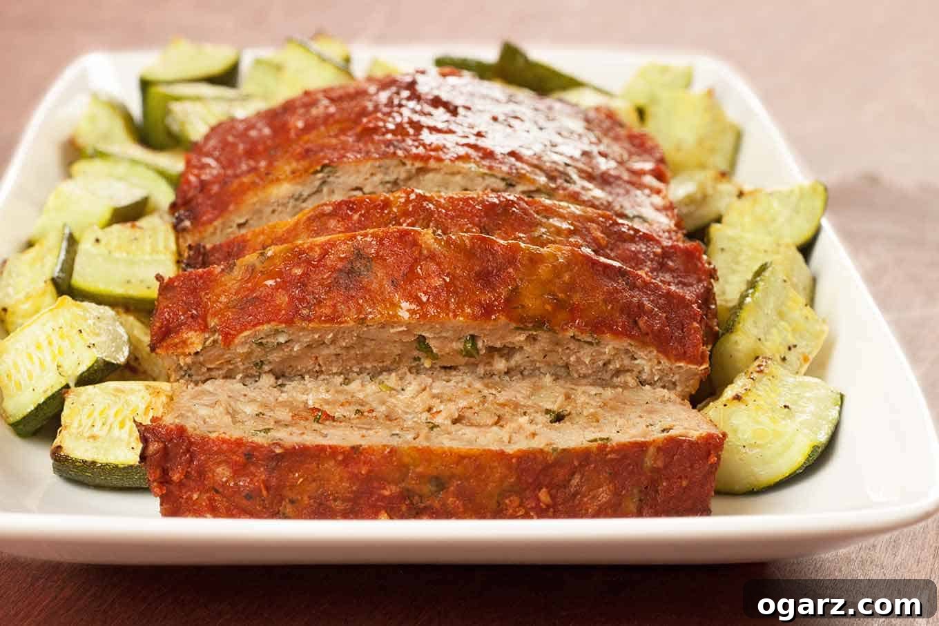 Tuscan Turkey Sausage Meatloaf 3 A white, oblong serving dish with thick slices of balsamic glazed turkey meatloaf and roasted zucchini on a brown wooden table.