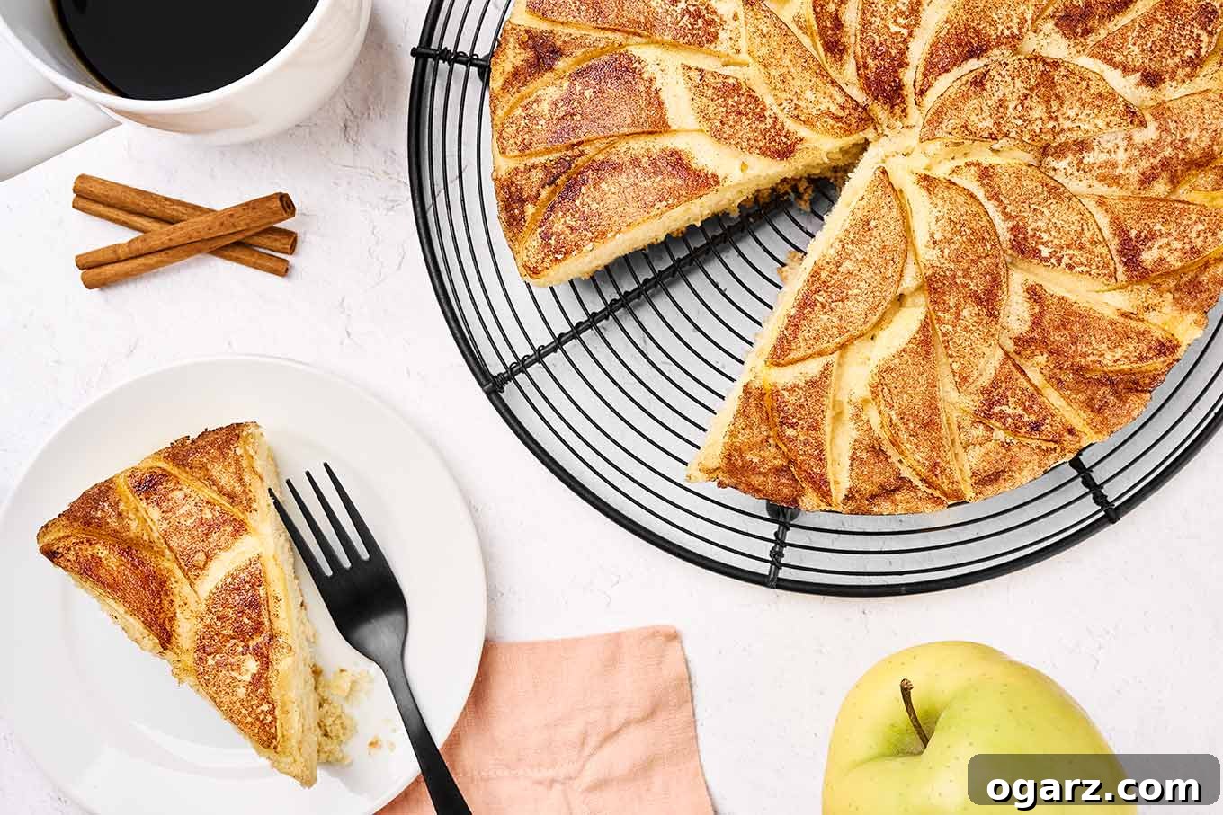 A slice of apple cake on a plate with a black fork, a cup of coffee in the corner, and the rest of the cake on a black wire cooling rack.