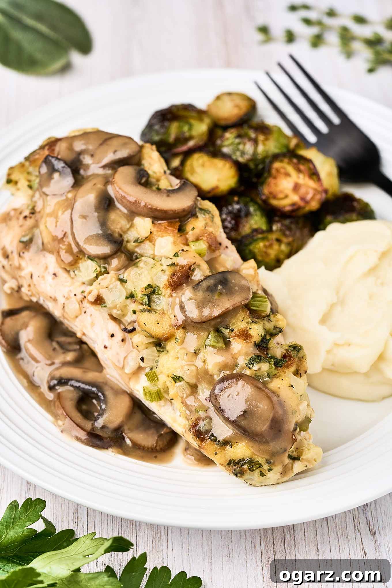 Succulent Baked Turkey Tenderloins with Savory Stuffing 2 A white plate with a stuffed turkey tenderloin topped with mushroom gravy, sides dishes of roasted Brussels sprouts and mashed potatoes, and a black fork.