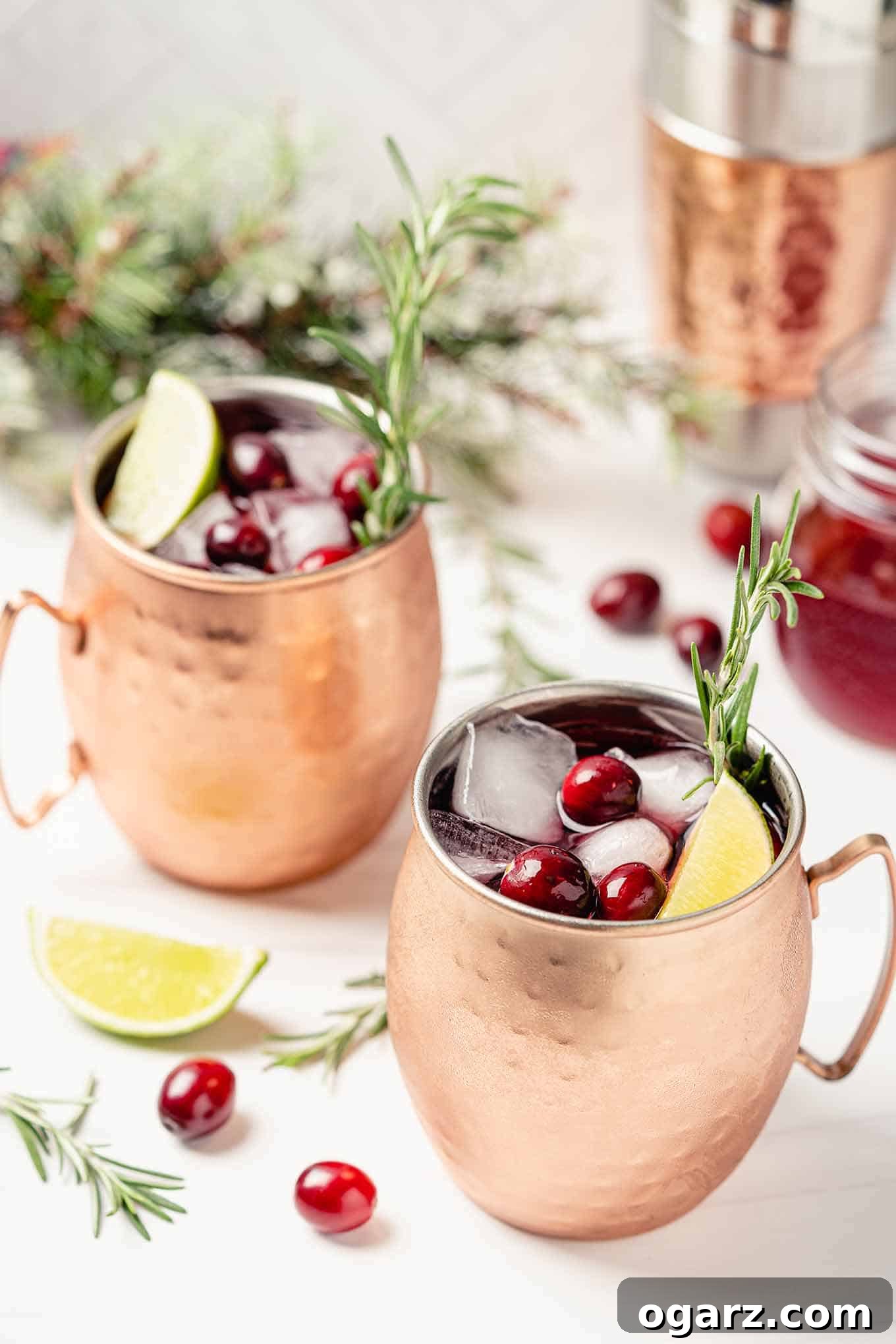 Crimson Mule 2 Two glistening copper mugs brimming with festive cranberry Moscow mules, elegantly garnished with fresh cranberries, zesty lime wheels, and aromatic rosemary sprigs. The scene evokes a perfect holiday toast.
