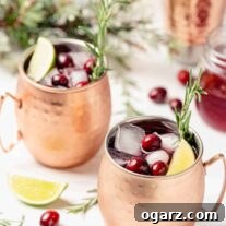 Two copper mugs filled with Moscow Mule cocktail, along with ice cubes, fresh cranberries, lime wedges and sprigs of fresh rosemary.