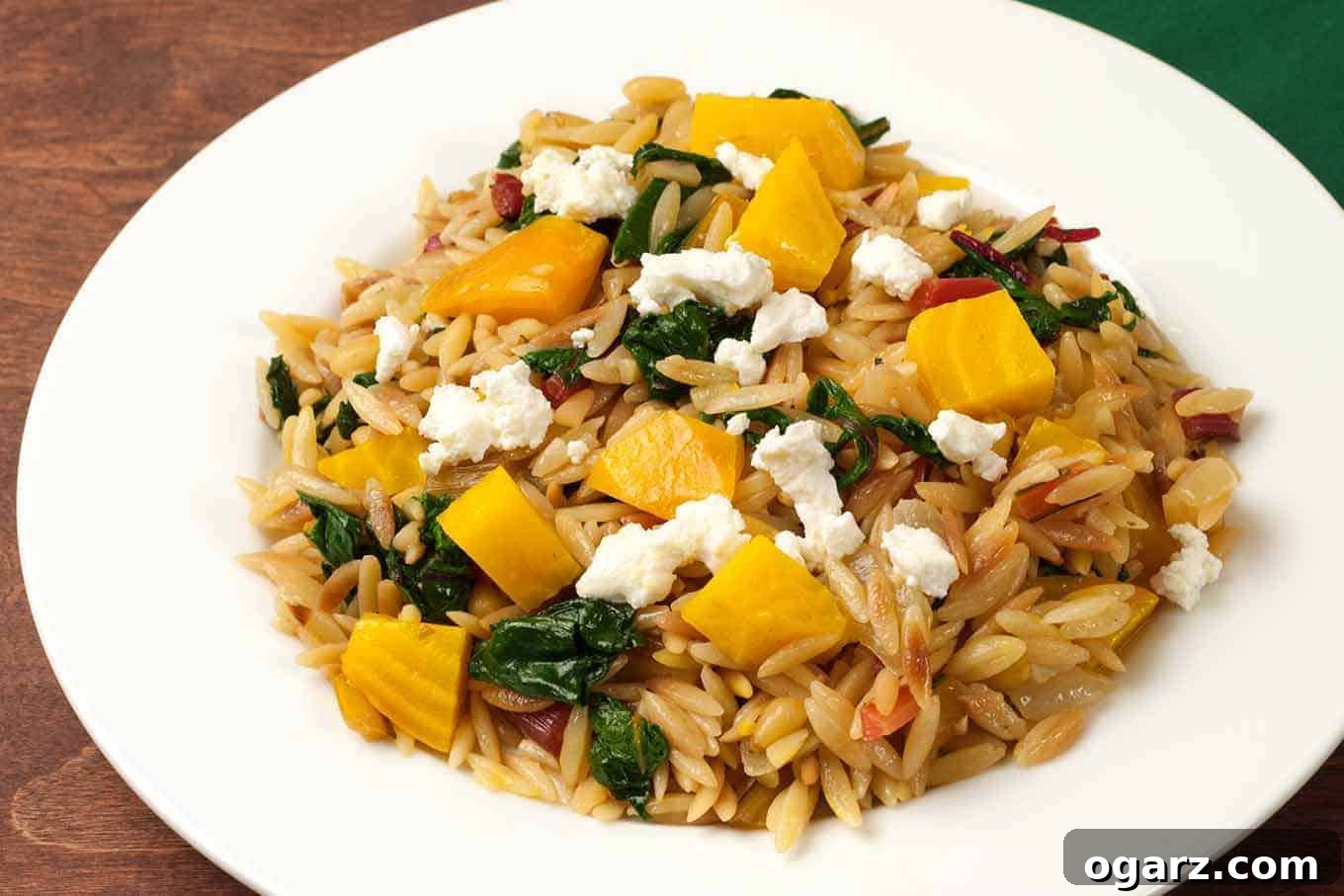A vibrant plate of orzo pasta featuring tender roasted golden beets, sautéed Swiss chard and beet greens, all elegantly topped with crumbled goat cheese and a sprinkle of pine nuts.