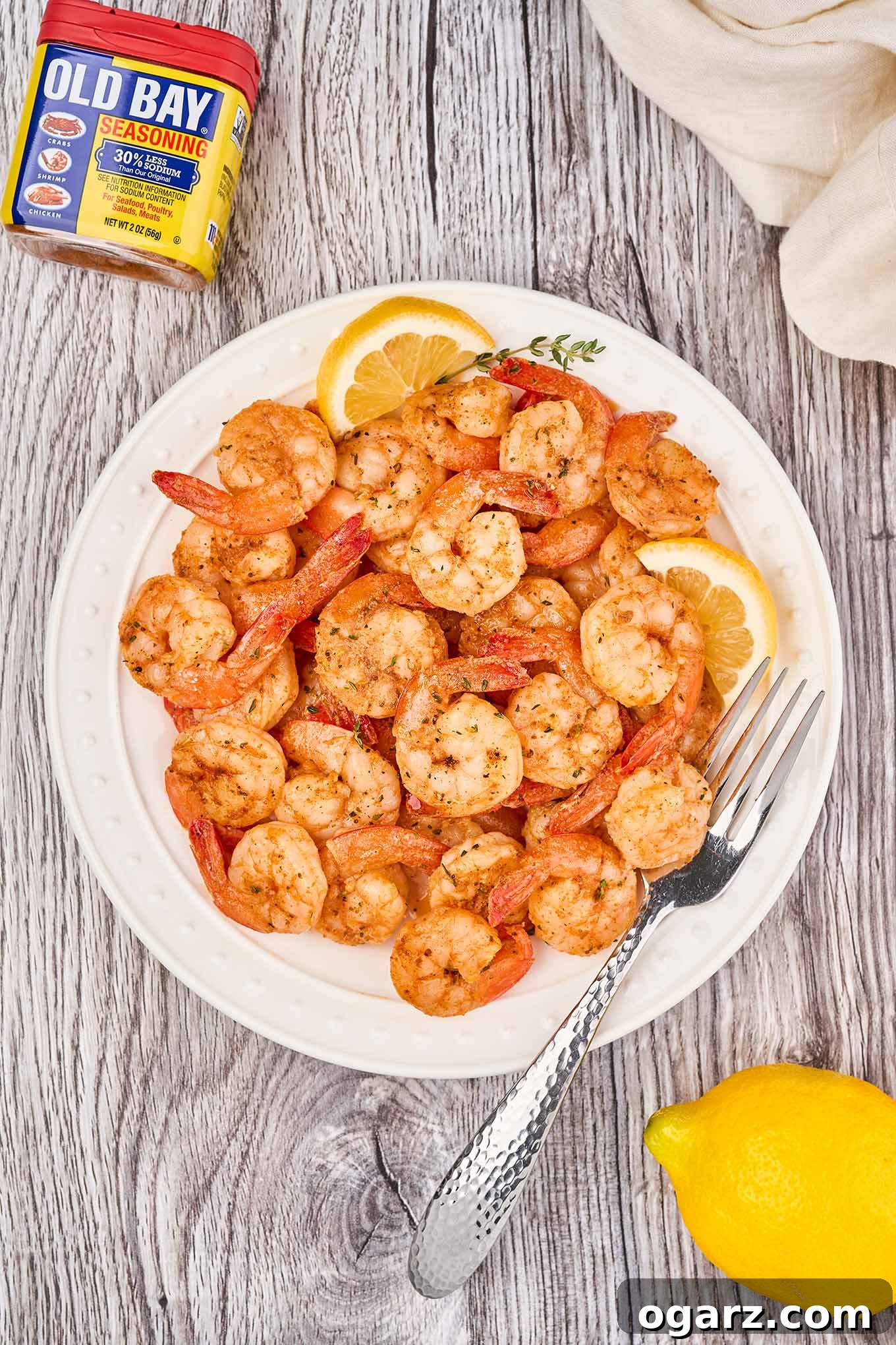 Zesty Old Bay Skillet Shrimp 4 A white plate piled with Old Bay sautéed shrimp and a fork, lemon wedges, a sprig of thyme, linen napkin, and Old Bay tin in the background.