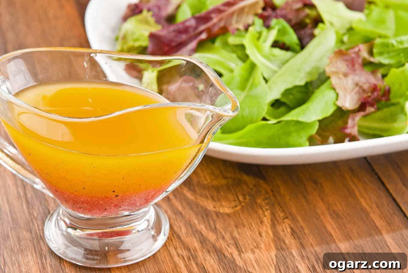 Essential Homemade Salad Dressings 3 A glass gravy boat filled with vinaigrette dressing in front of a plate of fresh mixed greens on a wooden table.
