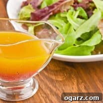 A glass gravy boat filled with vinaigrette dressing in front of a plate of fresh mixed greens on a wooden table.
