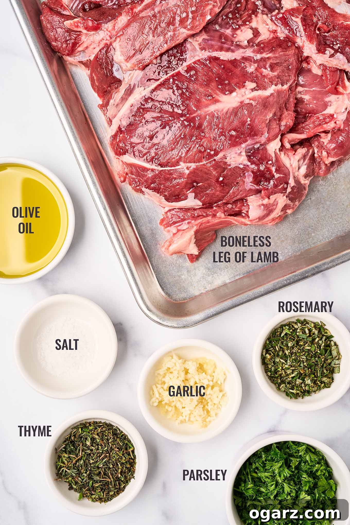Succulent Roasted Boneless Lamb Leg 3 A labeled, overhead view of the ingredients for roasted boneless leg of lamb with garlic-herb rub; boneless lamb leg, olive oil, salt, garlic, chopped fresh thyme, rosemary, and parsley.