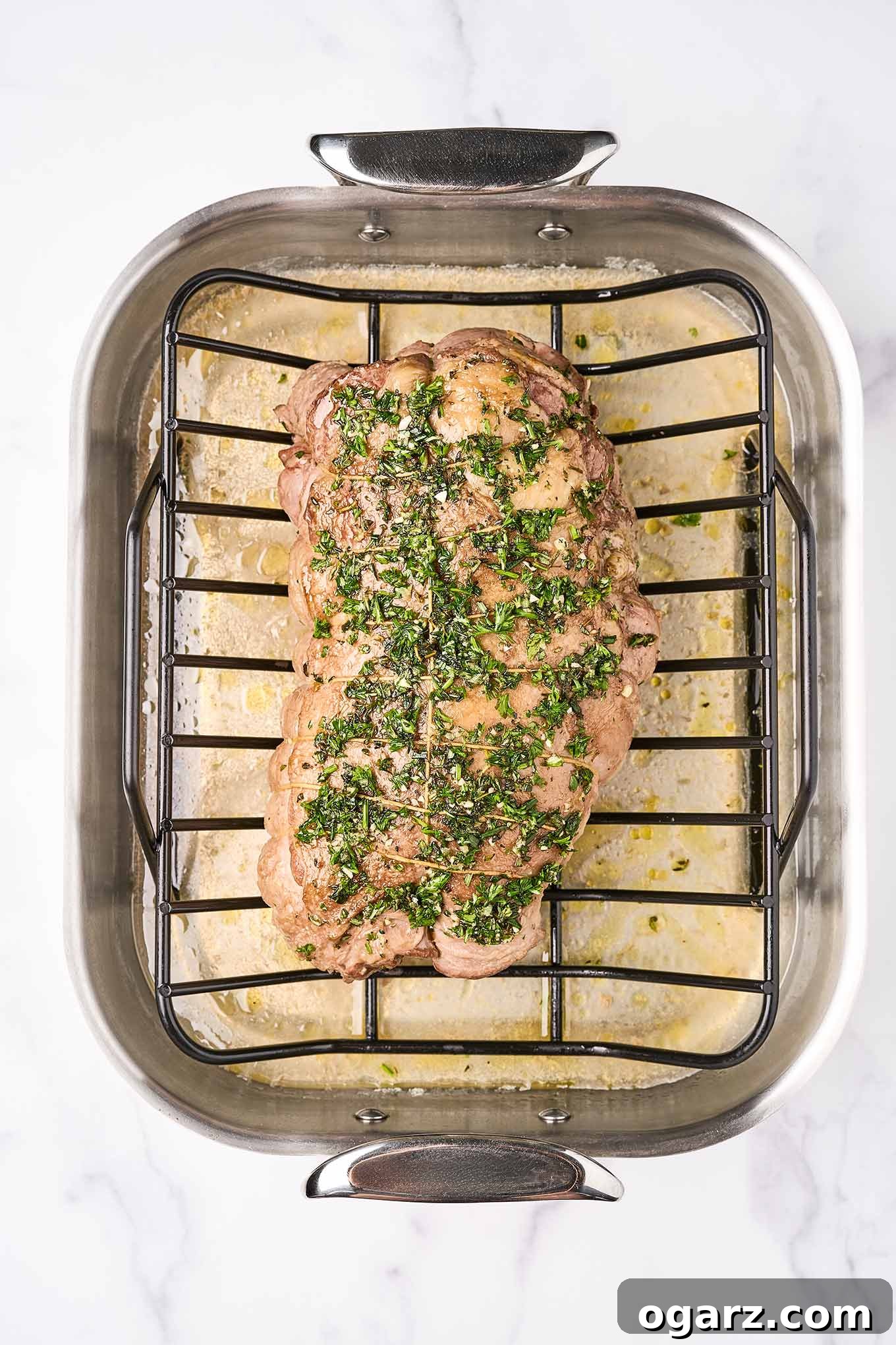 Succulent Roasted Boneless Lamb Leg 7 An overhead view of boneless leg of lamb in a roasting pan after having been in a hot oven for initial fat rendering, garlic and herb rub now applied on the top.