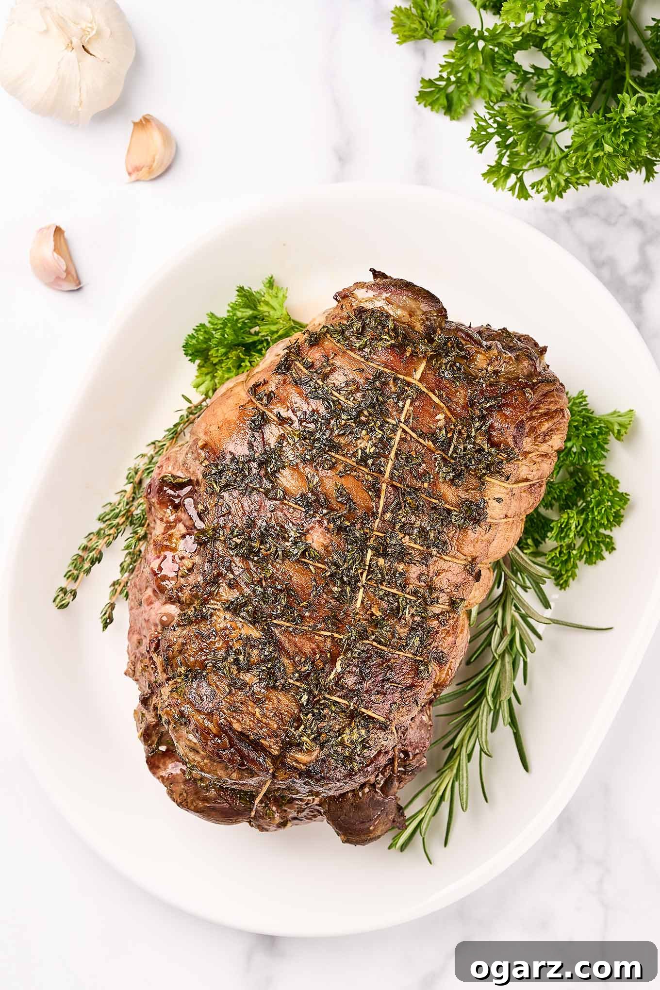 Succulent Roasted Boneless Lamb Leg 8 An overhead view of roasted boneless leg of lamb on a white oval platter with herb garnish, surrounded by sprigs of parsley, a bulb of garlic, a few scattered cloves.