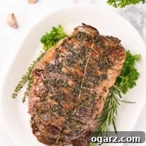 Succulent Roasted Boneless Lamb Leg 9 An overhead view of roasted boneless leg of lamb on a white oval platter with herb garnish, surrounded by sprigs of parsley, a bulb of garlic, a few scattered cloves.