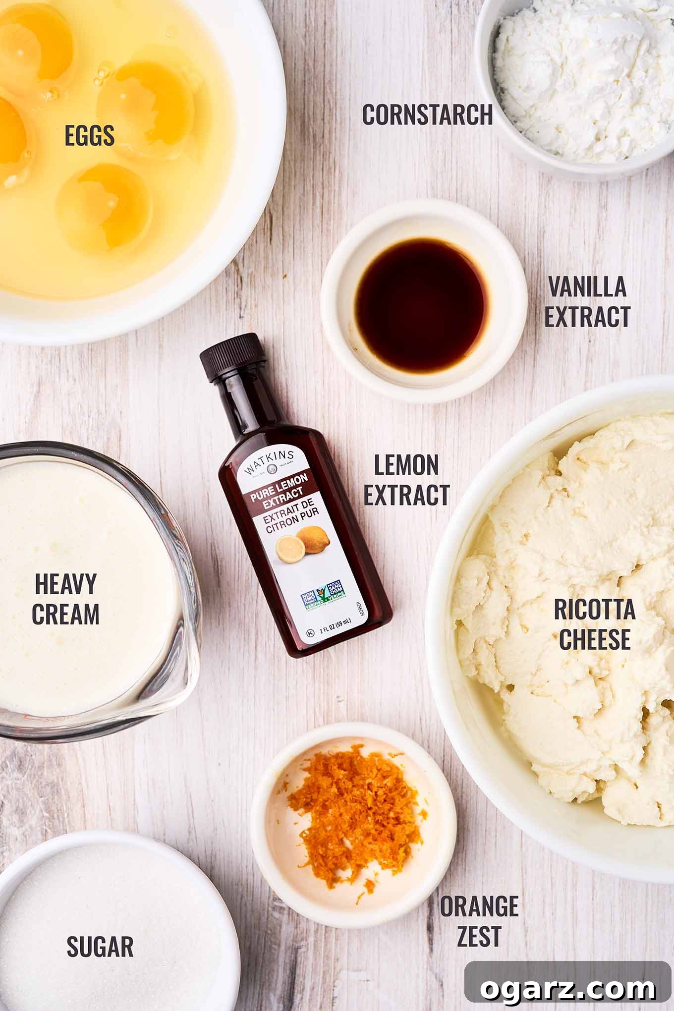 A labeled, overhead view of the ingredients for ricotta pie filling; eggs, cornstarch, heavy cream, ricotta cheese, lemon and vanilla extracts, orange zest, and sugar.