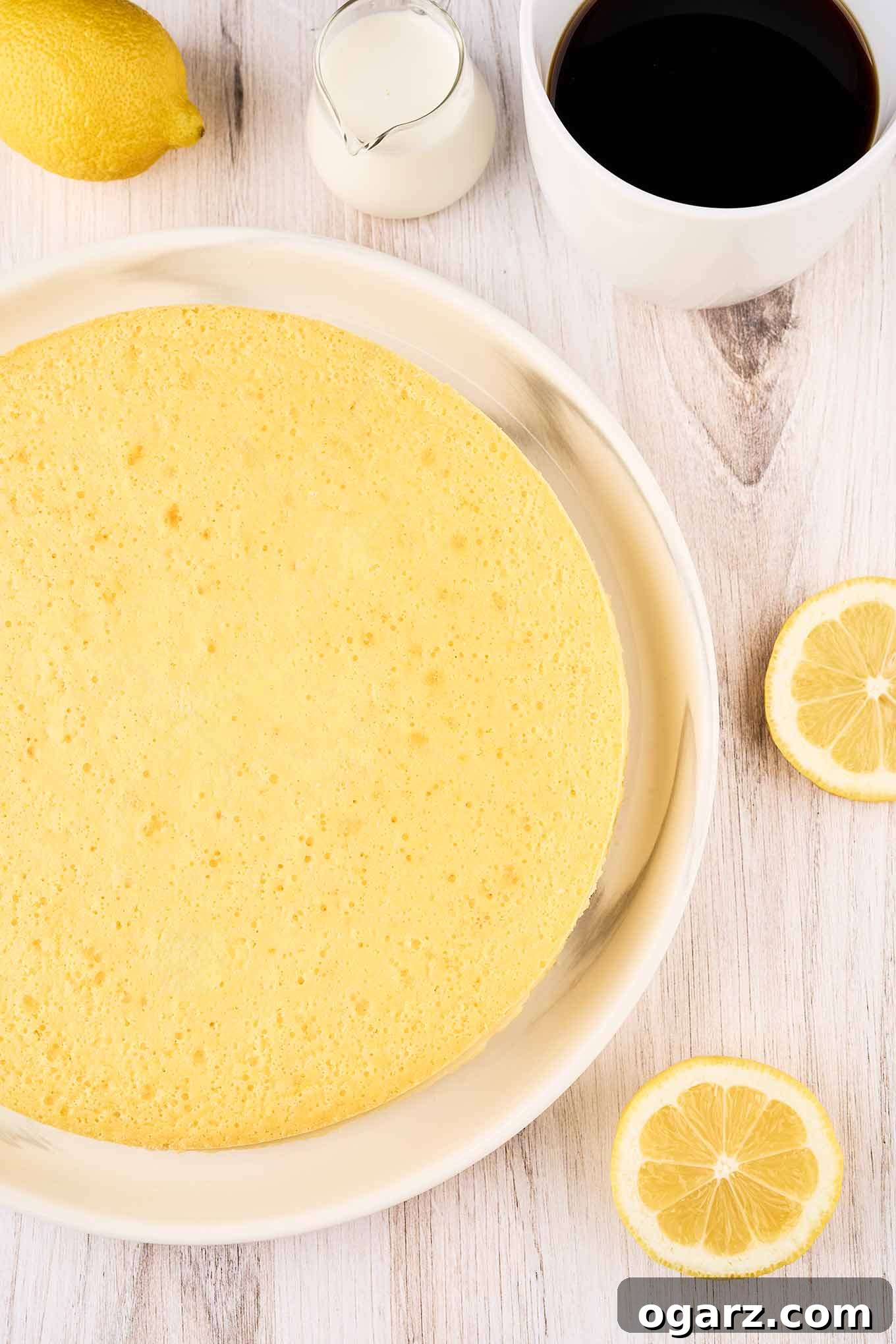 An overhead view of freshly baked ricotta pie on a platter surrounded by a cup of coffee, a pitcher of cream, a whole lemon, and lemon slices.