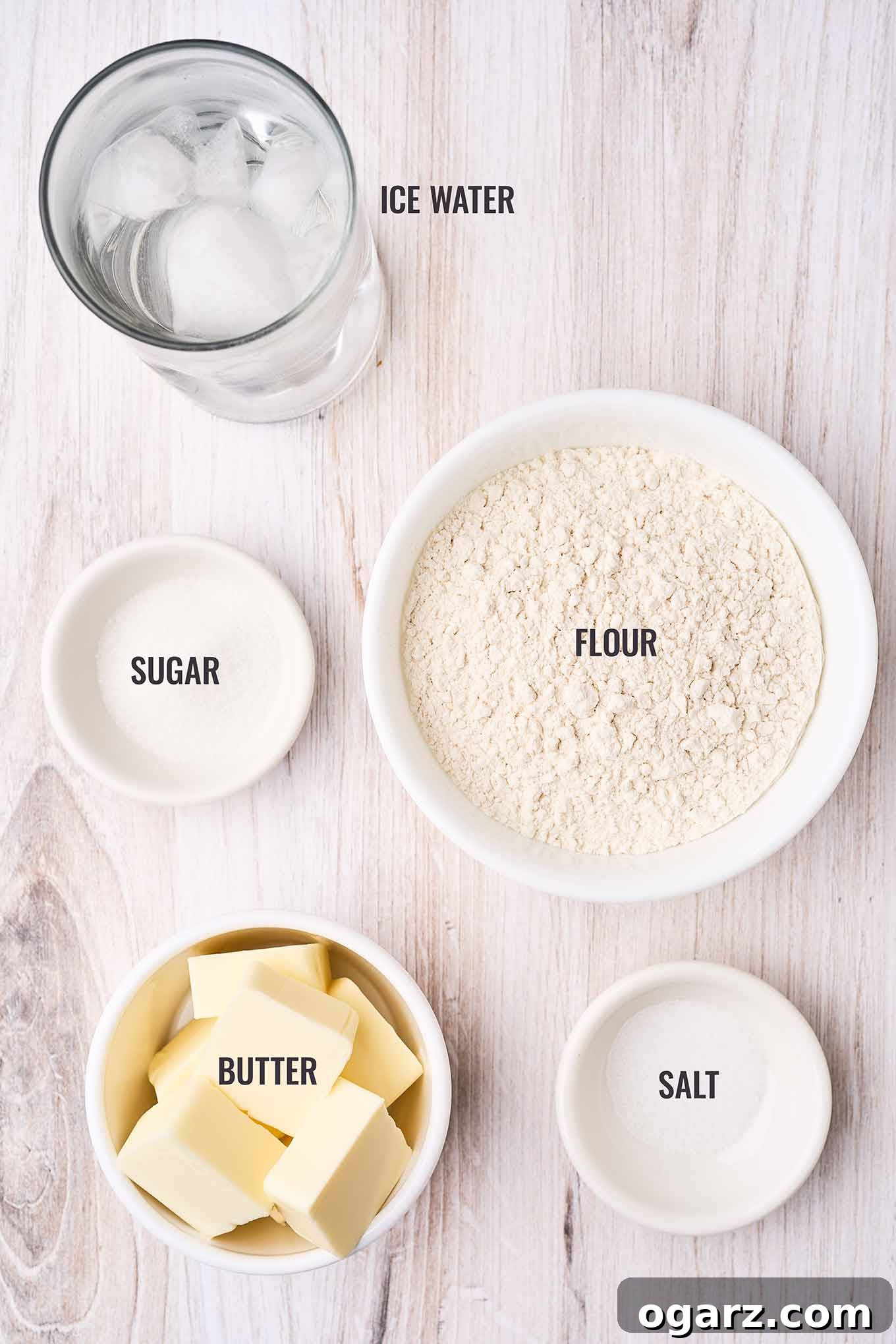 A labeled, overhead view of the ingredients for ricotta pie crust; flour, sugar, unsalted butter, salt, and ice water.