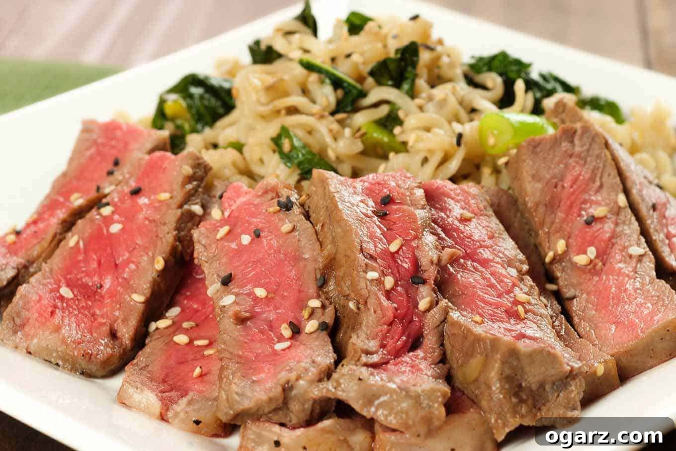 Sliced mirin-marinated strip steak topped with sesame seeds, served alongside scallion and kale ramen noodles on a white plate.