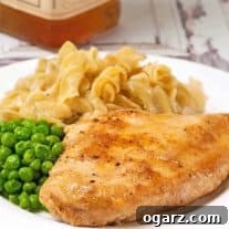 Whiskey glazed chicken breast served with buttered egg noodles and green peas on a white plate.