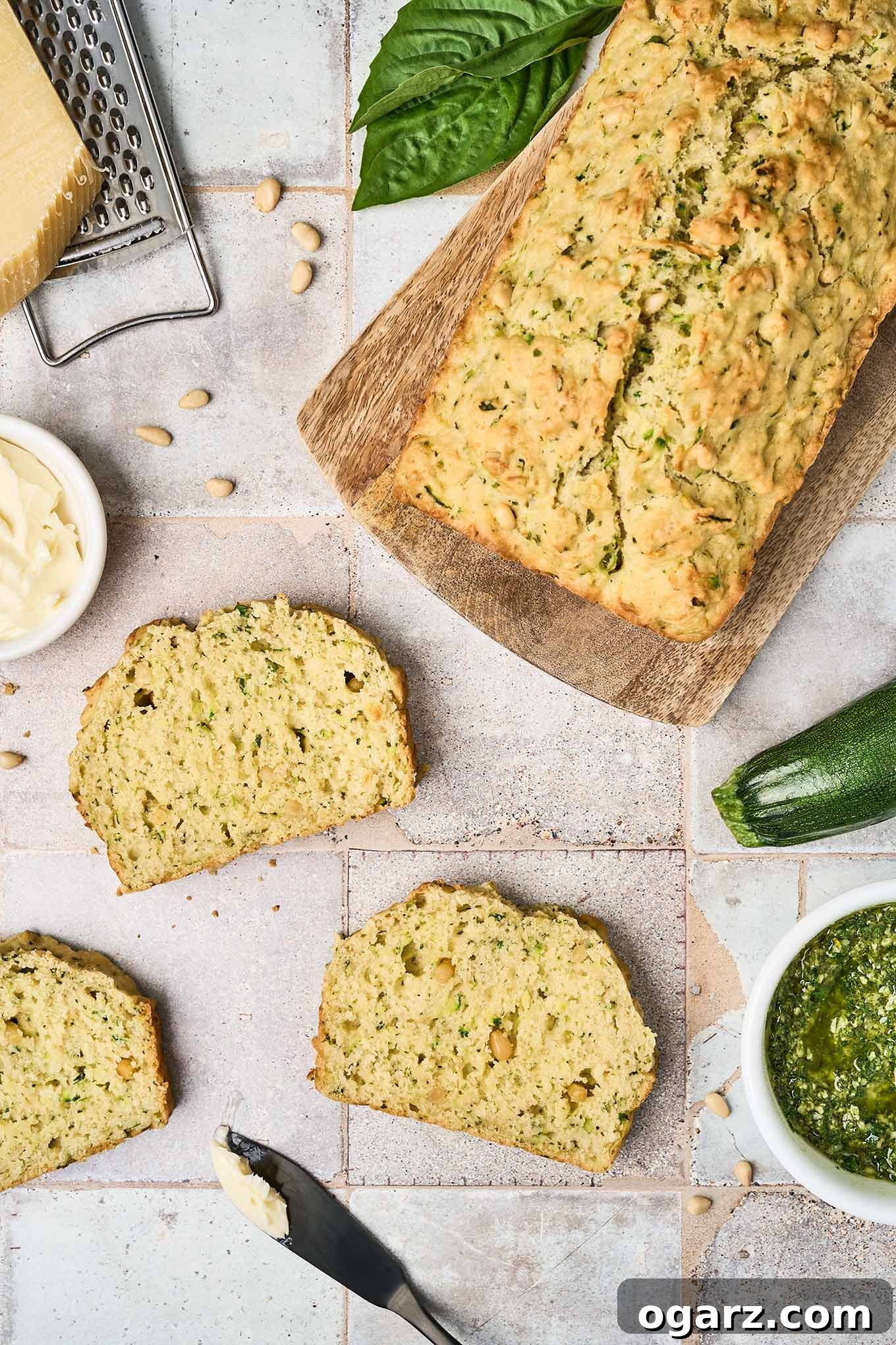 Herbed Zucchini Loaf 2 Three slices of savory pesto zucchini bread on a tile surface surrounded by a cutting board with the remainder of the loaf on it, a whole zucchini, a bowl of pesto, a dish of butter, Parmesan and a grater, pine nuts and basil leaves.