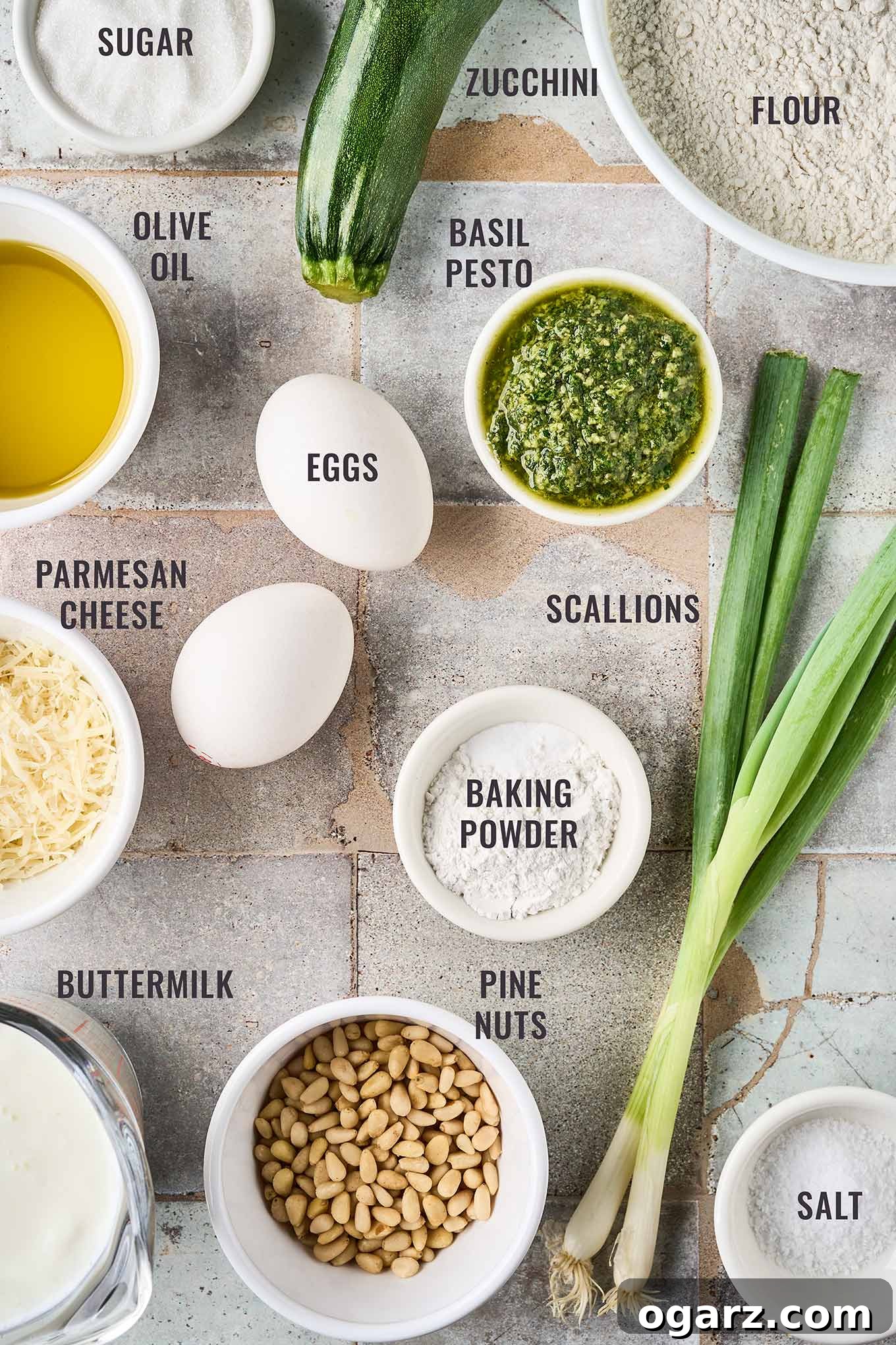 Herbed Zucchini Loaf 3 A labeled, overhead photo of the ingredients needed for savory pesto zucchini bread; flour, eggs, baking powder, basil pesto, scallions, pine nuts, buttermilk, Parmesan cheese, and olive oil.