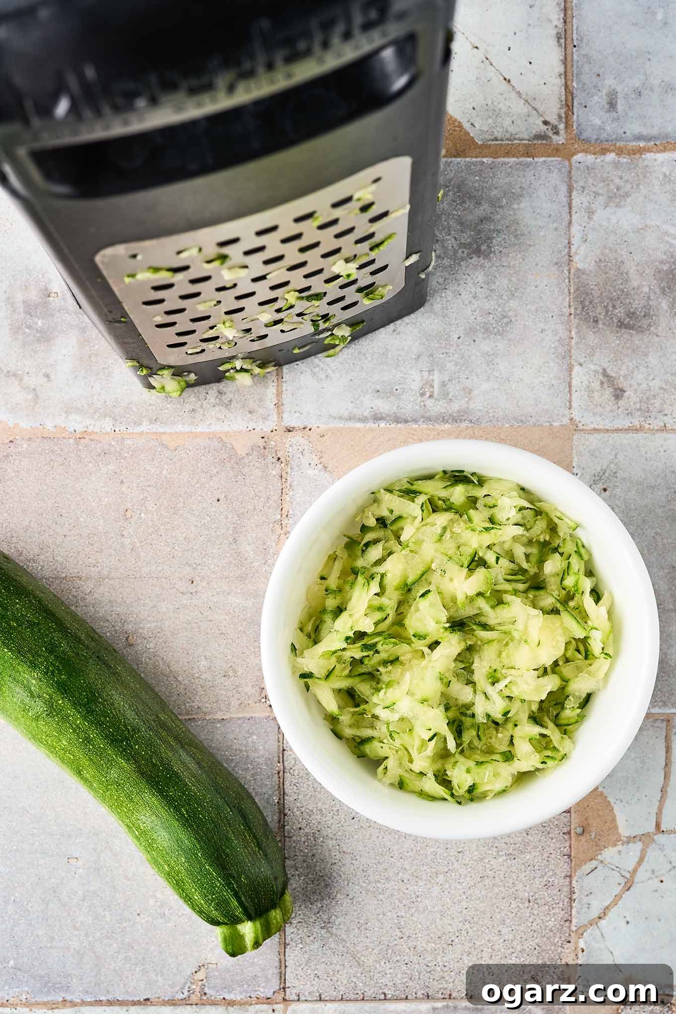 Herbed Zucchini Loaf 4 Coarsely grated zucchini in a bowl next to a box grater showing the large holes, a whole zucchini to the side.