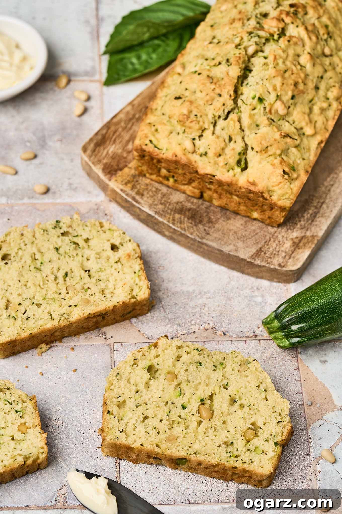 Herbed Zucchini Loaf 8 A close view of slices of savory zucchini bread on a tile countertop, a knife with butter on it, and a cutting board with a loaf of zucchini bread on it.