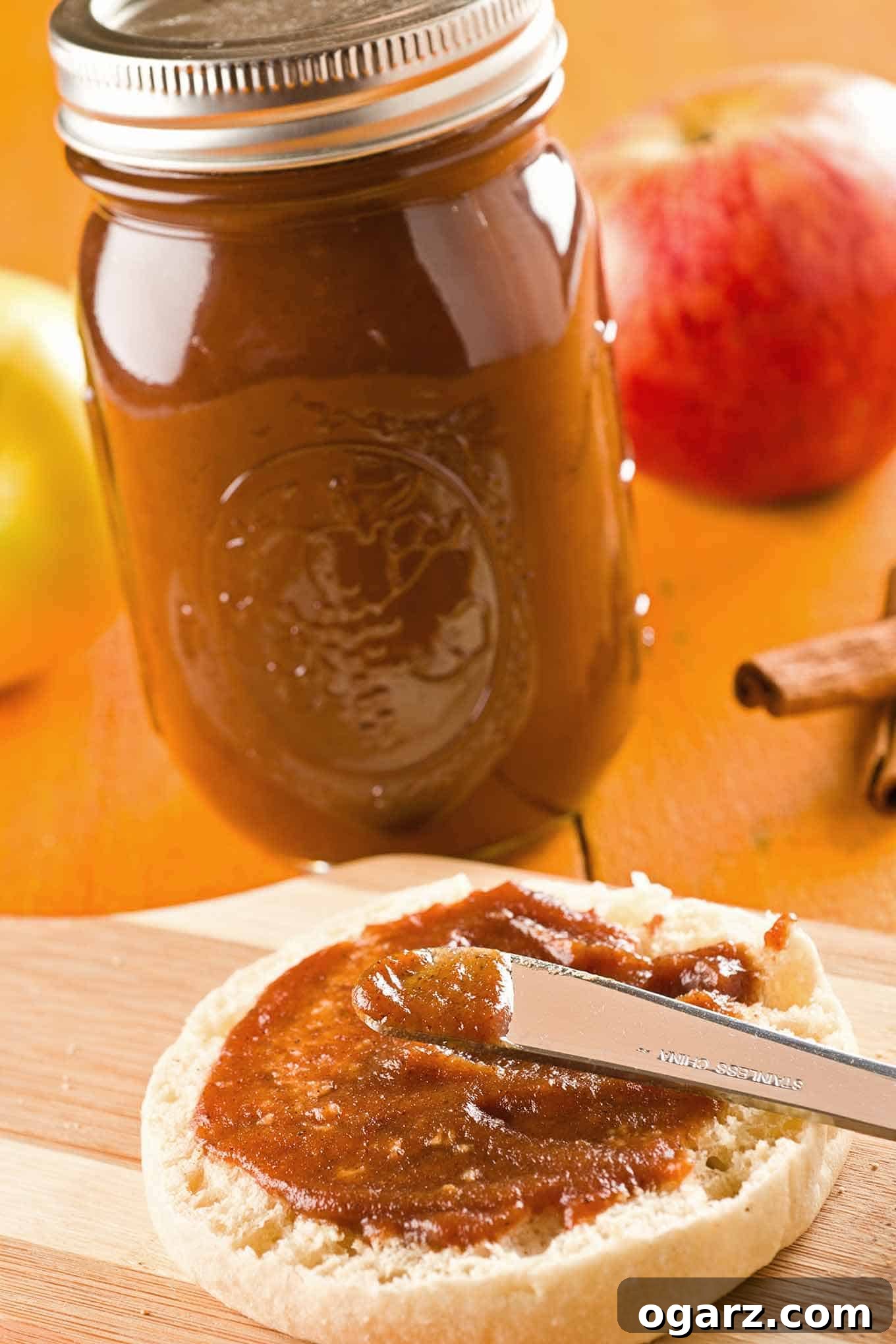 Velvety Slow Cooker Apple Butter 2 Delicious slow cooker apple butter being spread on an English muffin, with a jar of homemade apple butter, fresh apples, and cinnamon sticks in the background, showcasing its rich texture and warm colors.