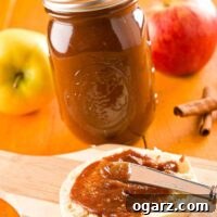 A close-up shot of homemade slow cooker apple butter being spread onto a toasted English muffin, with a full Mason jar of the butter and fresh apples in the background.