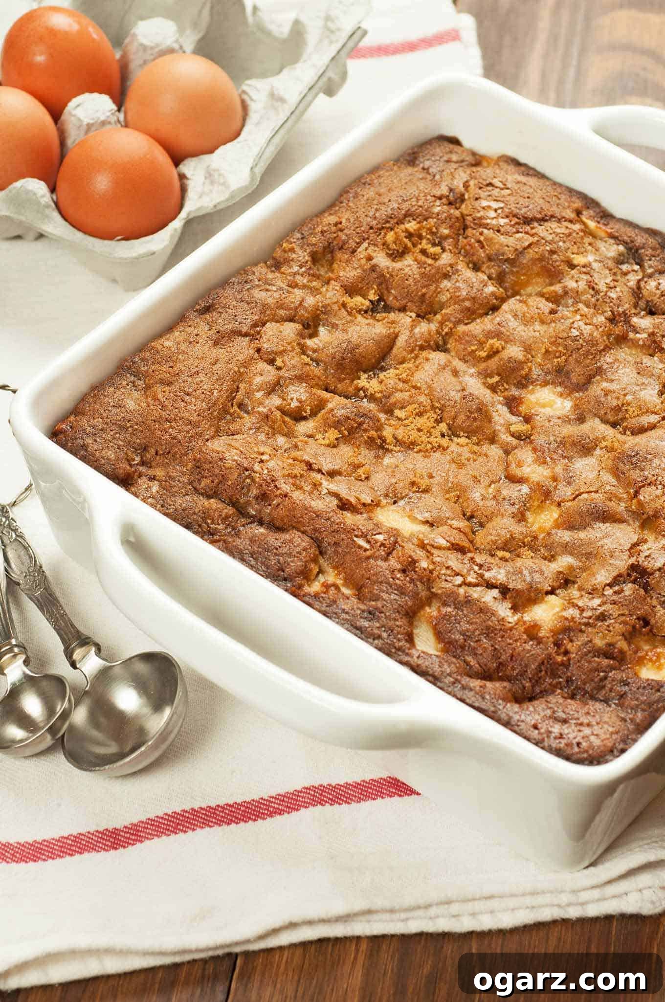 Freshly baked rum-spiced apple pudding cake in a white ceramic baking dish, displayed on a white towel with red stripe, with a carton of brown eggs and silver serving spoons nearby