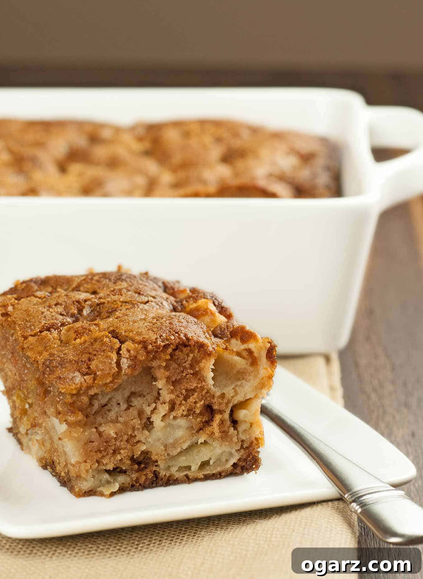 A single serving of spiced apple pudding cake on a plate with the whole cake in the background.