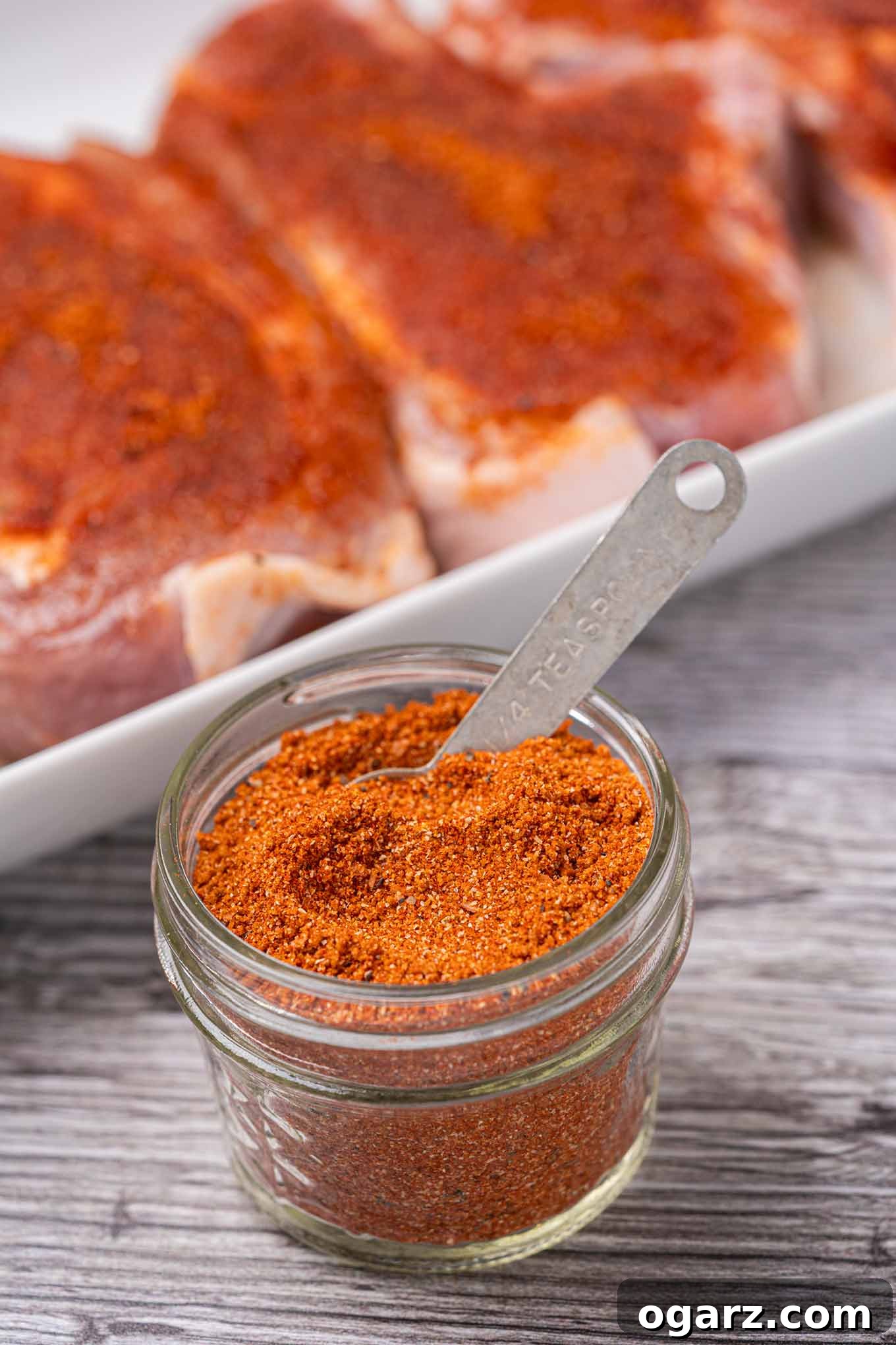 Effortless Grill Master Rub 2 A glass jar of BBQ spice rub with a measuring spoon in it with rub-coated pork chops in the background.
