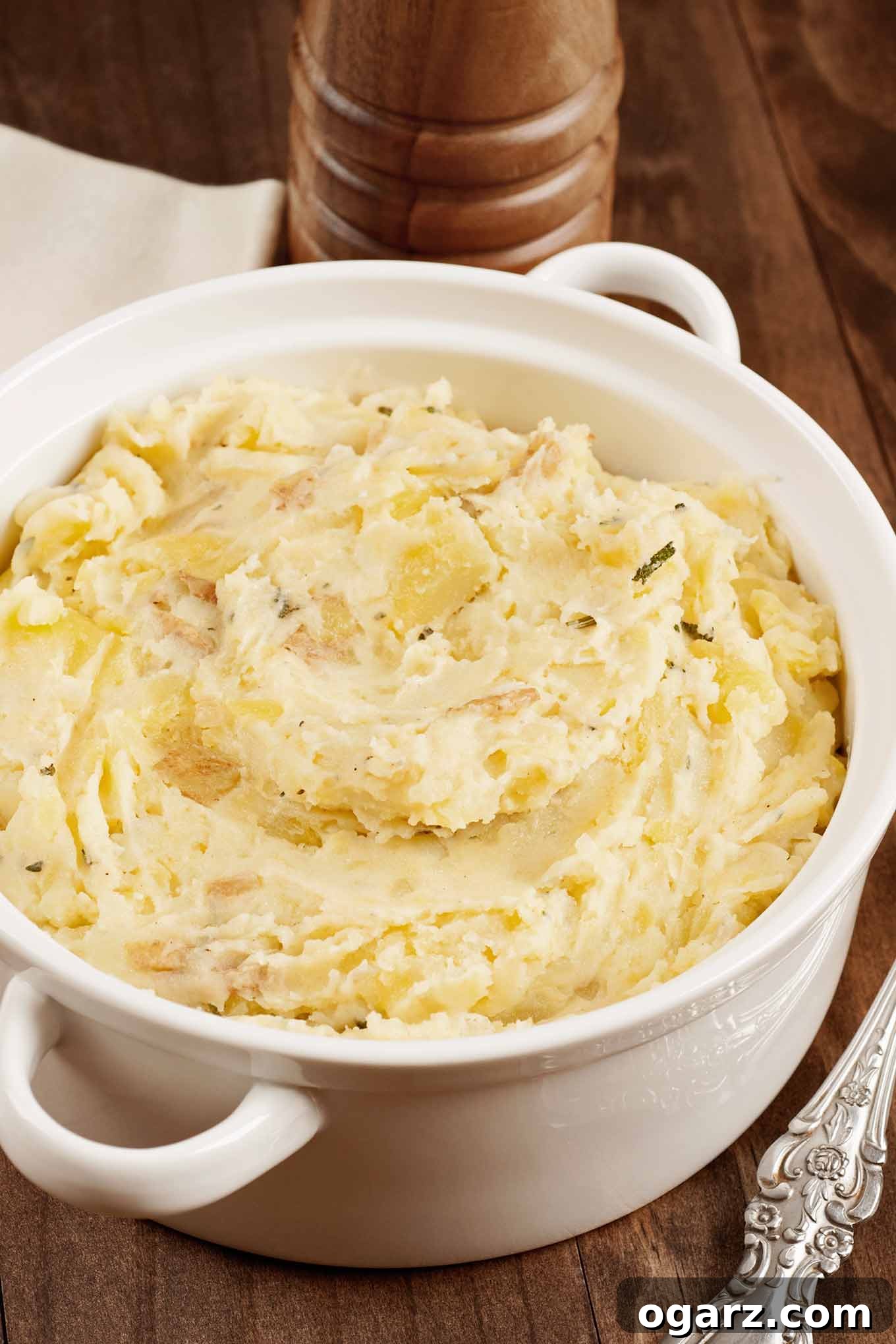 Hearty Potato Mash 2 A steaming bowl of rustic mashed potatoes, generously hand-mashed, infused with savory shallots and vibrant fresh herbs. A serving spoon rests invitingly to the side, with a classic wooden peppermill in the background, suggesting a wholesome, comforting meal.
