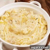 A round white casserole filled with rustic, hand-mashed potatoes with shallots and fresh herbs, serving spoon to the right, wooden peppermill to the rear.