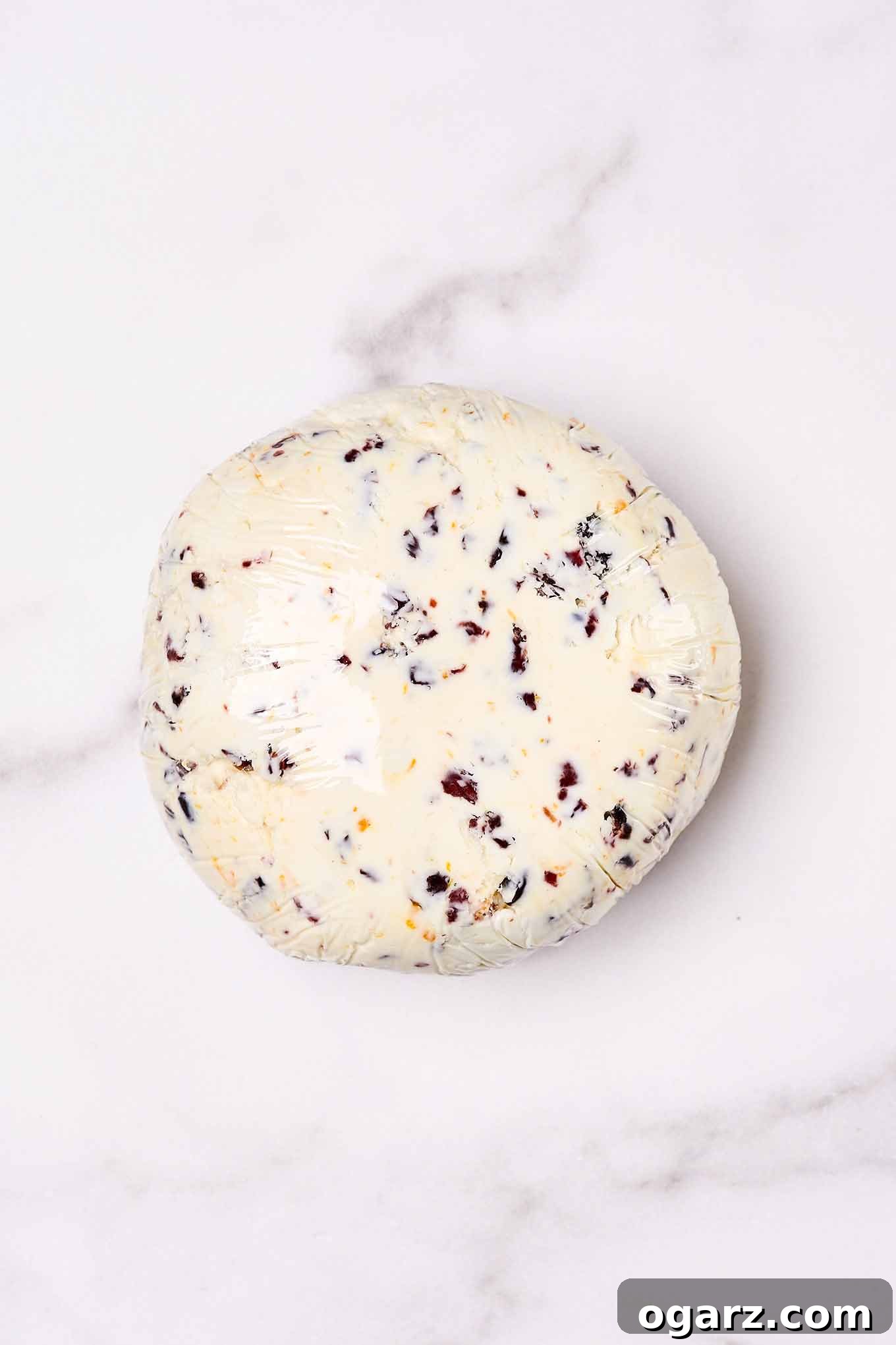 A cranberry cheese ball tightly wrapped in clear plastic wrap, ready for chilling on a sleek marble board, ensuring its shape is set.