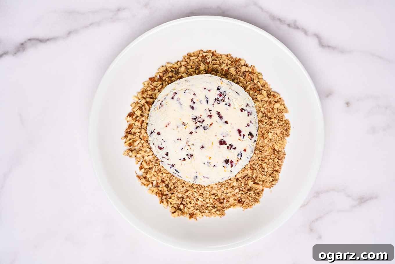 A festive cranberry cheese ball resting on a bed of finely chopped, toasted pecans on a white plate, poised for rolling to achieve a perfect, crunchy coating.