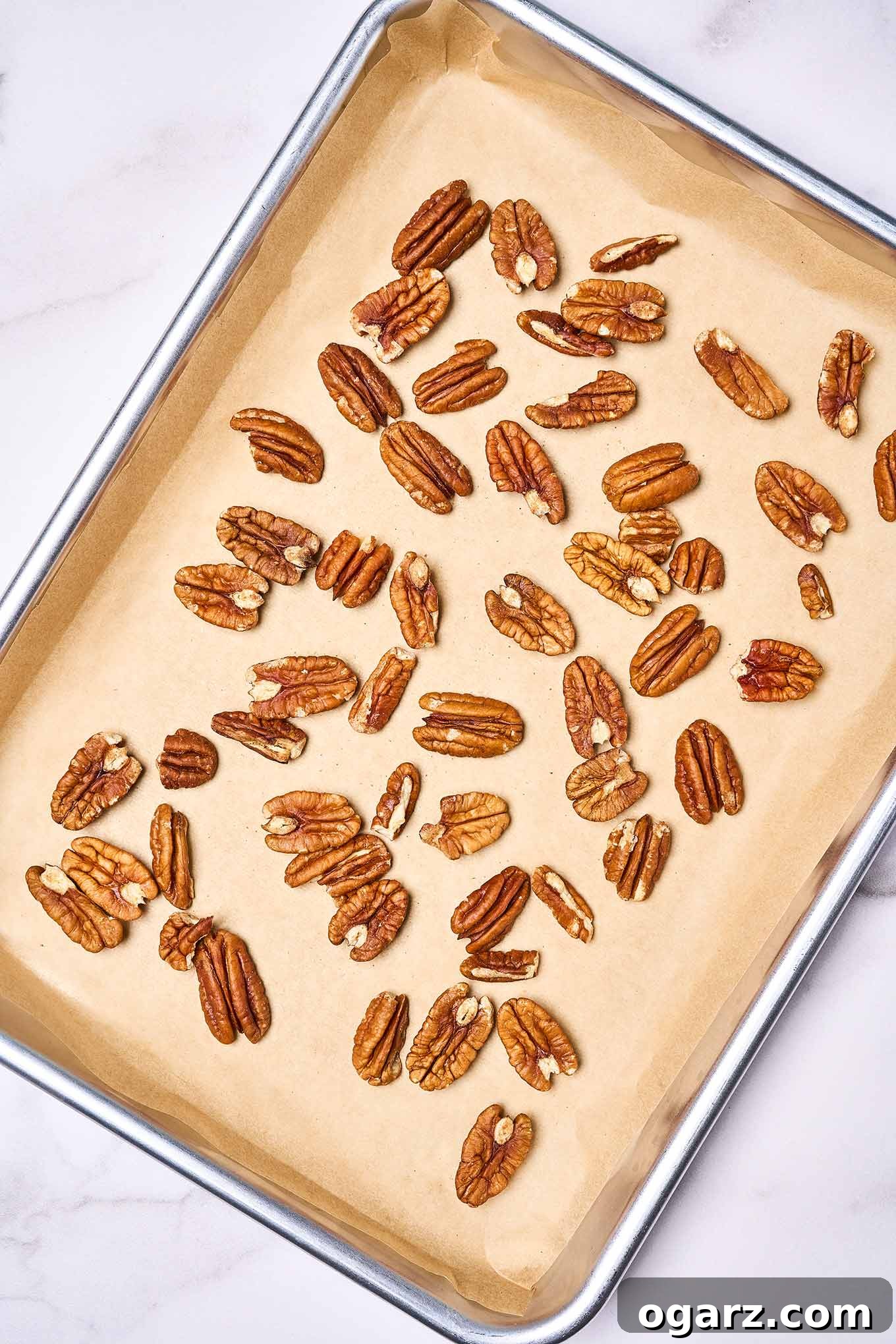 Pecan halves are meticulously spread in a single layer on a parchment-lined baking sheet, prepared for toasting in the oven to achieve a perfect golden brown.