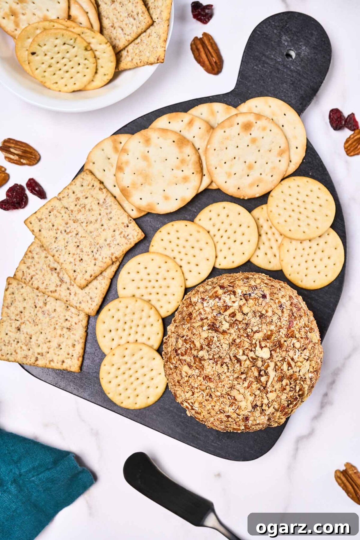 An overhead view of a beautifully presented cranberry cheese ball and an assortment of crackers arranged on a sleek black serving board, with more crackers, toasted pecans, and a spreader artfully placed around the board, inviting guests to indulge.