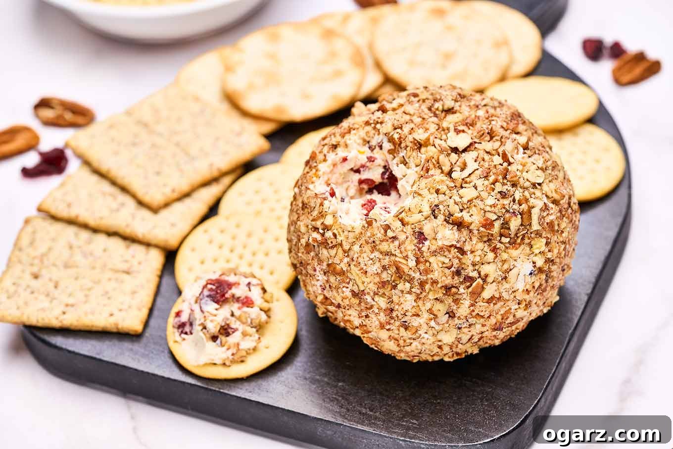 A black serving board showcasing a variety of crackers alongside a vibrant cranberry cheese ball, generously rolled in toasted nuts. A small portion of the cheese ball has been elegantly spread onto a cracker, tempting guests to savor its flavors.