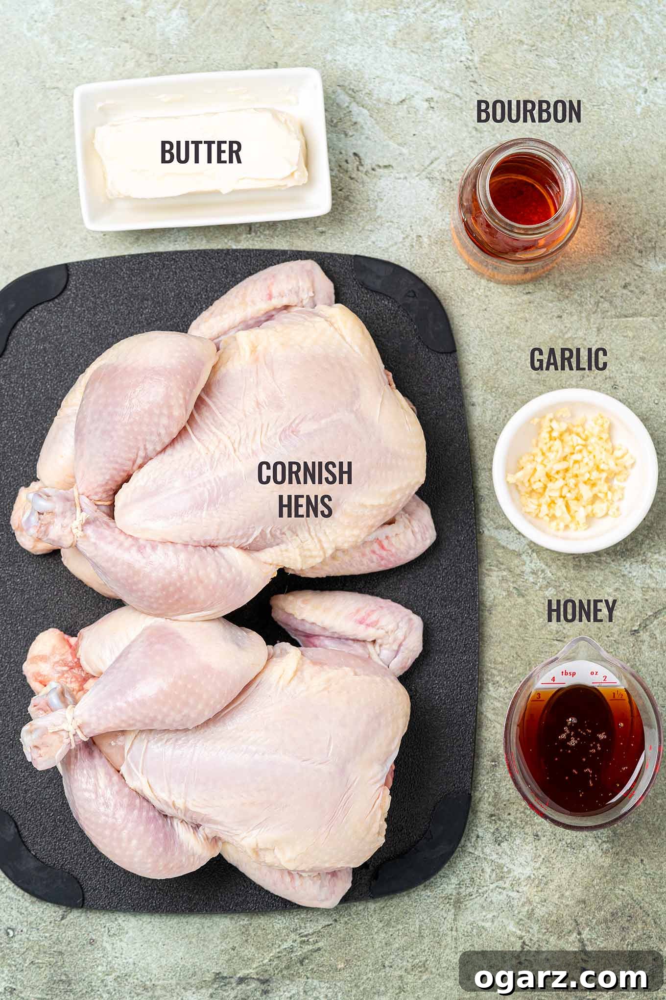 A labeled, overhead photo of the ingredients needed to make Cornish hens with bourbon glaze, including two cornish hens, butter, bourbon, garlic, and honey.