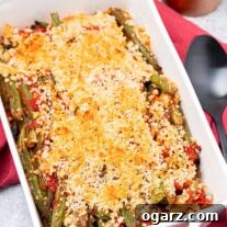 Fresh from the oven, a casserole of baked Italian-style green beans and tomatoes with crisp Parmesan-panko topping, a pepper mill and serving spoon in the background.