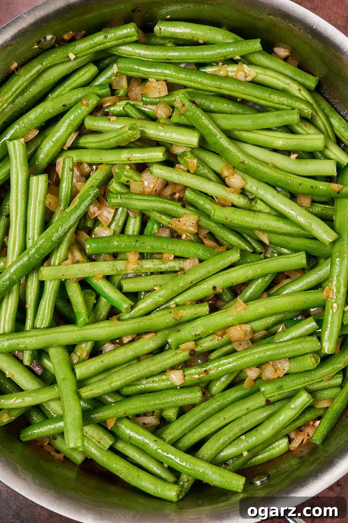 Trimmed, raw green beans in a sauté pan with sautéed onion and garlic, ready to be braised in chicken broth.