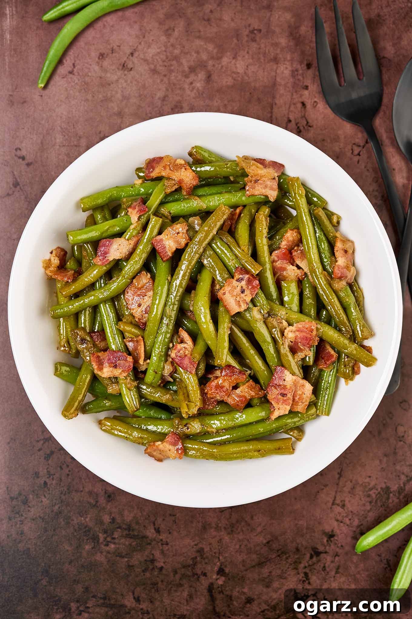 A round, white serving bowl of Southern style green beans with bacon on a rustic brown surface with a black serving fork to the right.